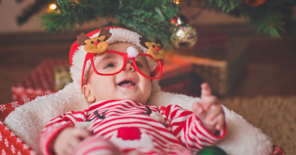Leende bebis med julsmyckade glasögon ligger i ett paket under en julgran.